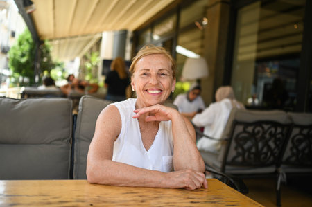 Happy senior woman with perfect smile and teeth sitting outdoors at summer sidewalk street cafe or...