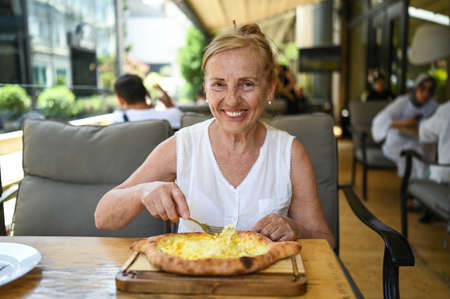 Happy senior woman with perfect smile eating adjarian khachapuri outdoors at summer sidewalk street cafe or restaurant terrace. Retired mature people holiday vacation, active lifestyle conceptの写真素材