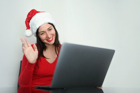 Beautiful smiling brunette woman in New Years Santa Claus hat works with laptop. Christmas and New Year office concept. Young female freelancer dressed all red in happy moodの写真素材