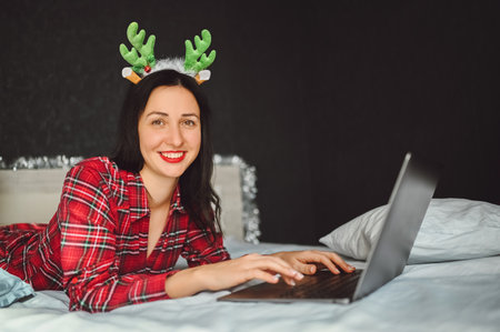 Beautiful excited smiling brunette woman in red pajamas lies in bed in Christmas reindeer horns works with laptop in dark interior bedroom. Young female freelancer at home in happy New Year mood.の写真素材