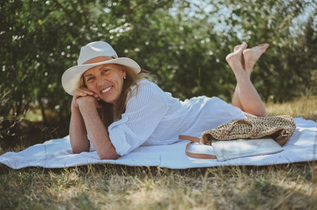 Senior happy woman laying on picnic blanket with laptop in bag at summer outdoors. Old elegant lady in straw hat on grass at countryside. Active retired people conceptの写真素材
