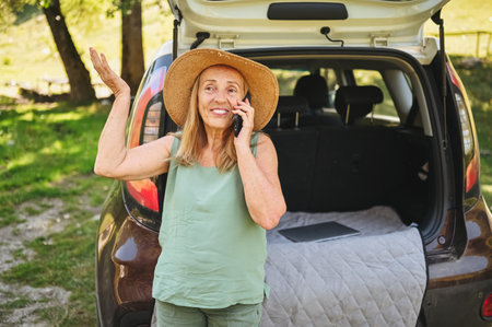 Senior active happy woman speaks phone car trunk or boot in camper sunny summer park - digital nomad freedom lifestyle concept, travel freelancer and vacations of retired old age people.の写真素材
