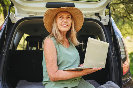 Senior happy woman working with laptop from the car trunk or boot in camper sunny summer park - digital nomad freedom lifestyle concept, travel freelancer and vacations of retired old age people.の写真素材