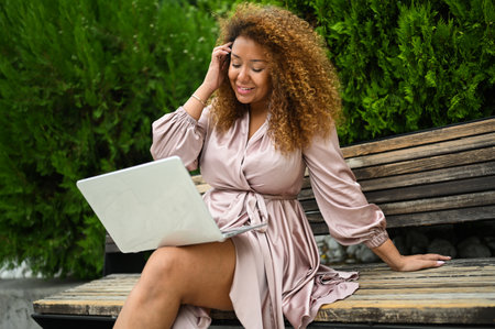 Attractive happy stylish plus size African American woman student freelancer afro hair studying online working on laptop computer outside at summer park. Diversity. Remote work, distance educationの写真素材