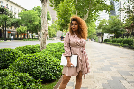 Attractive happy stylish plus size African American woman student freelancer afro hair with laptop computer outside, big urban city street at summer. Diversity. Remote office work, distance educationの写真素材