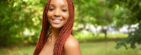 Close up portrait Beautiful young African American woman with red braids hair, perfect white teeth smiling outside, sunny summer day green foliage. Facial treatment. Cosmetology, skin care and spaの写真素材