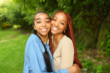 Close up street style portrait beautiful young African American female black couple. Braids hair, perfect white teeth smiling, sisters friends hugging walking outdoor in sunny summer day in green parkの写真素材