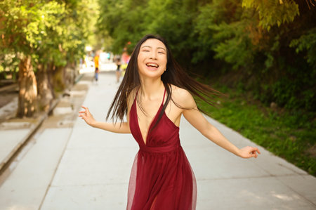 Street style outdoor portrait of Beautiful young Chinese Asian happy smiling woman walking outside sunny summer day. Attractive woman in red dress enjoying posing in green parkの写真素材