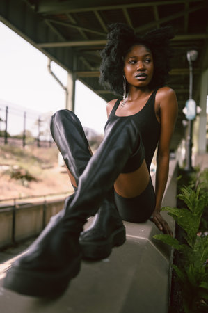 Fashion outdoor street style portrait Beautiful young African American woman posing outside on urban city landscape summer day wearing total black high leather boots.の写真素材
