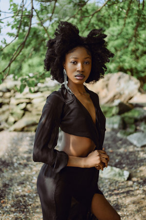Fashion outdoor beach portrait Beautiful young African American Black woman posing outside on sandy coast with rocks and green trees on sunny summer day. Cosmetology, natural skin body care and spaの写真素材