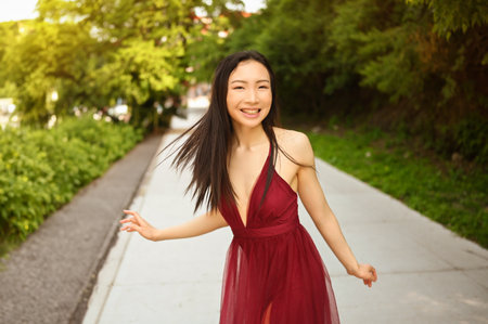 Street style outdoor portrait of Beautiful young Chinese Asian happy smiling woman walking outside sunny summer day. Attractive woman in red dress enjoying posing in green parkの写真素材
