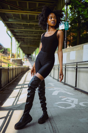 Fashion outdoor street style portrait Beautiful young African American woman posing outside on urban city landscape summer day wearing total black high leather boots.の写真素材