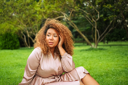 Close up portrait of Young beautiful African American woman enjoys a walk in summer public green park. A happy woman plus size wearing silk pink dress sitting resting on grassの写真素材