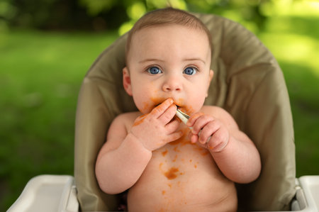 Cute adorable infant funny dirty baby trying to eat himself with fruit or vegetable puree smeared on...
