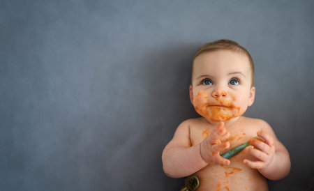 Cute adorable infant funny dirty baby trying to eat himself with fruit or vegetable puree smeared on...