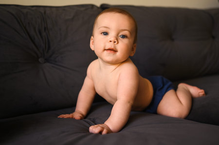 Cute emotional smiling funny infant boy sitting on straight arms on dark grey deep navy blue couch...
