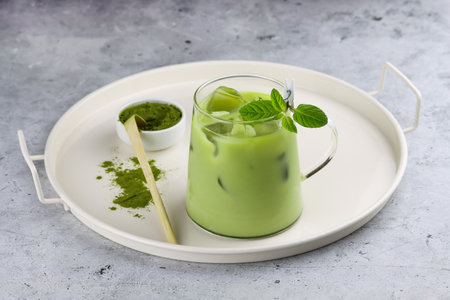 Antioxidant matcha drink made of ground powder of green tea leaves in transparent cup with milk, ice cubes and fresh mint on a serving tray with matcha spoon and matcha powder bowl. Iced matcha green teaの写真素材