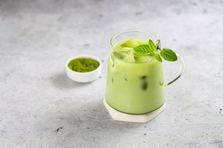 Antioxidant matcha drink made of ground powder of green tea leaves in transparent cup with milk, ice cubes and fresh mint on a serving tray with matcha spoon and matcha powder bowl. Iced matcha green tea with milkの写真素材