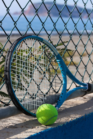 Tennis racket and tennis ball on the sandy beach in front of the sea and mountain on a sunny day. Summer sport concept. Tennis training by the sea. Tennis on the beach background. Workout at the sea shoreの写真素材