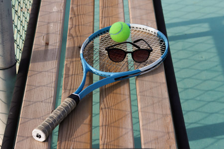 Tennis racket, tennis ball and sun glasses on a bench at the blue hard tennis court. Hard lighting. Summer sport concept. Active lifestyle and healthy living, close upの写真素材