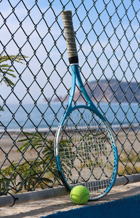 Tennis racket and tennis ball on the sandy beach in front of the sea and mountain on a sunny day. Summer sport concept. Tennis training by the sea. Tennis on the beach background. Workout at the seashoreの写真素材