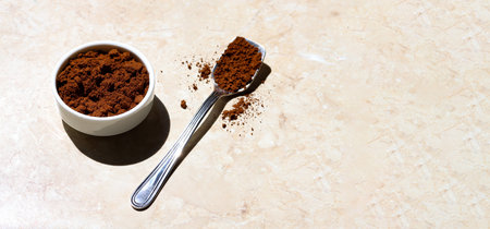 A ceramic white bowl and a silver spoon filled with ground coffee on a light marble table on a sunny...
