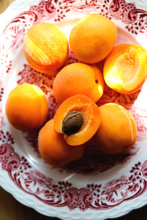 Delicious ripe apricots in a plate on the table on a sunny day, close-up, vertical. Pile of fresh whole juicy orange apricots and apricot halves with a seed on a plate of English porcelain, top view.の写真素材