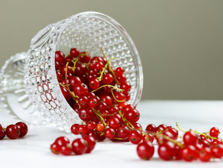 Fresh red currants fall out of a glass bowl on marble table. Ripe red currant berries in transparent glass on light background with copy space. Minimalistic photo of red currants, selective focusの写真素材