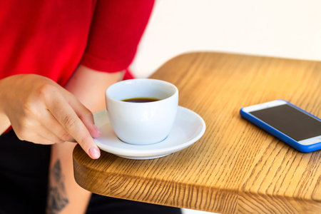 Image of woman's hand holding white ceramic cup with filter coffee, sitting on the wooden table in...