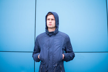 young guy in a jacket standing against a blue wall