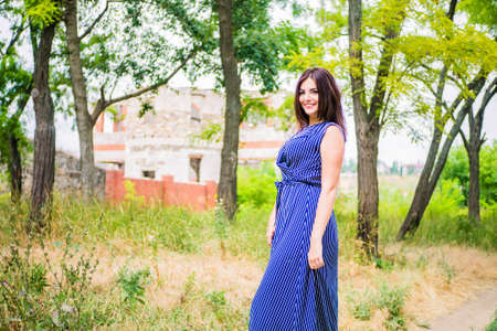 Girl brunette in a blue long dress on a dirt road against a background of trees and natureの写真素材