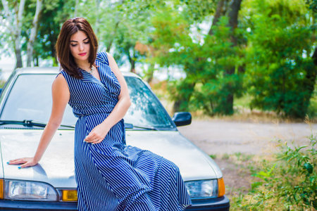 Brunette girl in a blue long dress sits on the hood of a carの写真素材