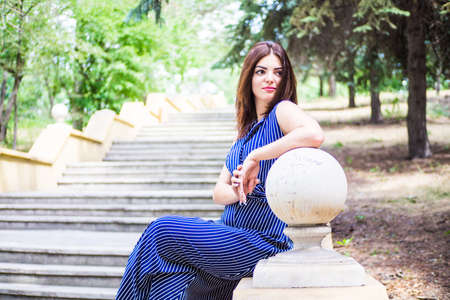 The brunette girl in a blue long dress is sitting in the park on the steps near the flashlight in the afternoonの写真素材