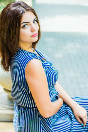 The brunette girl in a blue long dress is sitting in the park on the steps near the flashlight in the afternoonの写真素材