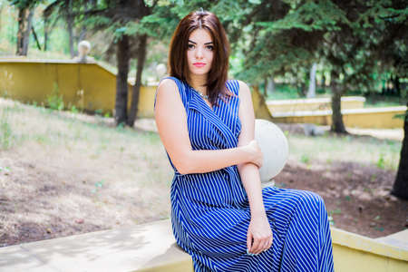 The brunette girl in a blue long dress is sitting in the park on the steps near the flashlight in the afternoonの写真素材