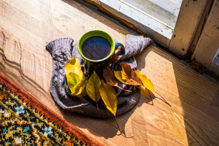 A black cup with coffee near the window, with yellow leaves and a knitted brown scarf on a wooden...