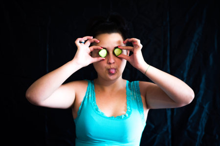 A young girl cares for her skin, putting blue clay on her face and fresh cucumbers in her eyes from...