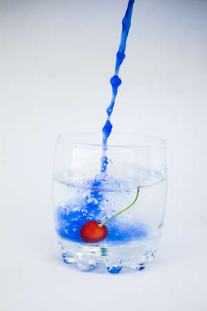 In a transparent glass with clean water, with cherries at the bottom, blue paint is poured in a dynamic movement and paints the water blue on a white background.の写真素材
