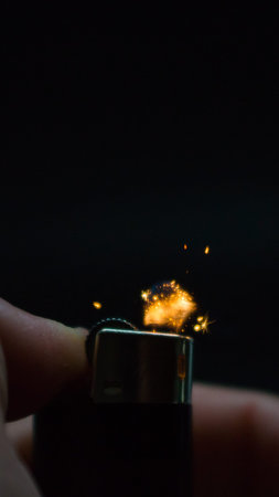 A person has lighters in his hands and he lights it through a flint wheel, the lighter ignites, sparks and fire appear, close-up on a black background.の写真素材