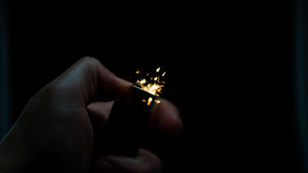 A person has lighters in his hands and he lights it through a flint wheel, the lighter ignites, sparks and fire appear, close-up on a black background.の写真素材