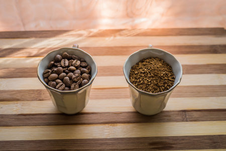 On a wooden plank there are two cups filled with whole roasted coffee beans and instant coffee...