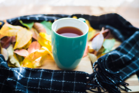 Hot flower tea in a blue mug on a wooden background, tea wrapped in a black scarf and scattered with autumn leaves, yellow, red, gold.の写真素材