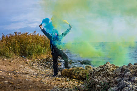 A boy twists blue and yellow smoke bombs in both hands on the seashore with a beautiful view of the horizon.の写真素材
