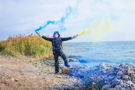 A boy twists blue and yellow smoke bombs in both hands on the seashore with a beautiful view of the...