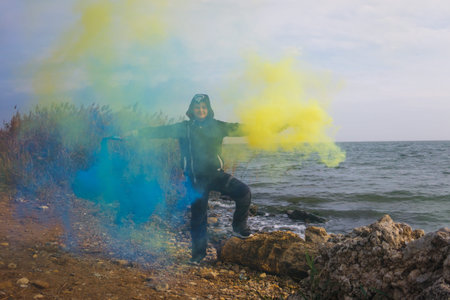 A boy twists blue and yellow smoke bombs in both hands on the seashore with a beautiful view of the...