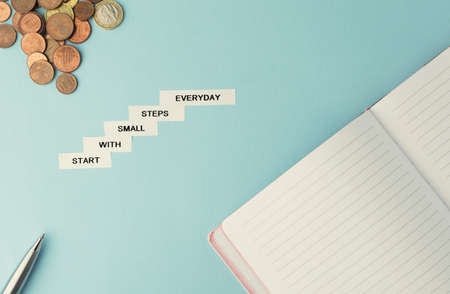 motivation business quote start with small steps everyday. White sticky notes on blue background with note book, pen and coins. Saving money and starting business. Planning, future.の写真素材