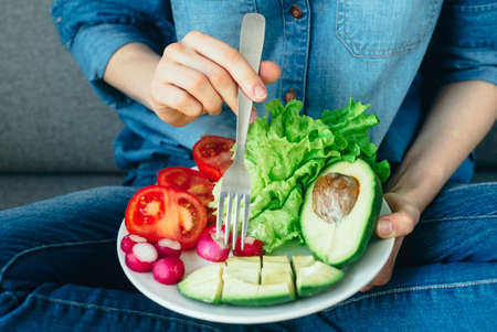 young woman eating fresh vegetables at home. Vegan eating avocado, salad, radish and tomatosの写真素材