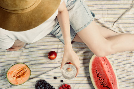summer picnic setting. Woman in jean shorts and hat eatining water melon, melon, blueberries,...