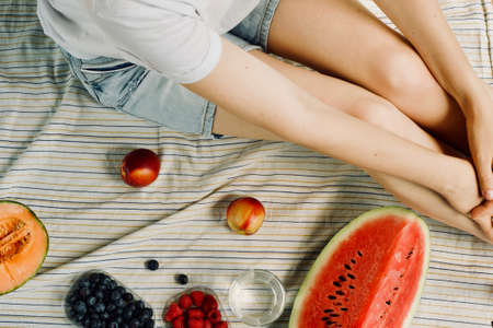 summer picnic setting. Woman in jean shorts and hat eatining water melon, melon, blueberries, raspberries and peaches, drinking water. Outdoor gathering concept.の写真素材