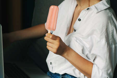 Young woman eating ice cream using laptop computer. Female working on laptop in an outdoor, sitting on sofa at homeの写真素材